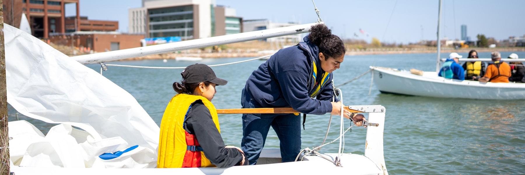 Two students work on sailboat in water near UMass Boston.