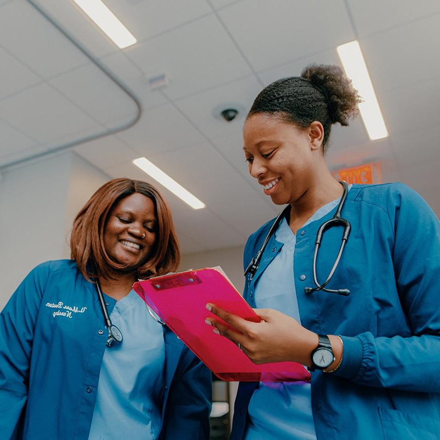 Nursing two students in scrubs hold clipboard in lab.